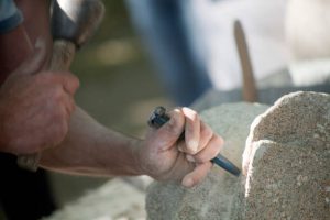 Close-up of craftsman's hands carving stone, holding chisel , hand moving hitting with hammer, during a traditional public stone-carving festival, outdoors, sunlight lighting up granite stone block, spanish culture. Copy space available on the right.